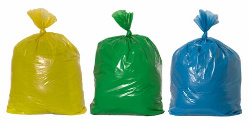 Three large plastic rubbish sacks are positioned upright on a paved surface, with the yellow bag on the left, the green bag in the middle, and the blue bag on the right. Each sack is tightly gathered and knotted at the top, exhibiting a glossy finish that reflects light. The yellow sack has a bright, vivid hue, while the green and blue sacks feature rich, distinct colours. The sacks appear to contain general waste, as indicated by their full and slightly bulging appearance. The background is plain and neutral, with no additional objects or environmental details visible, suggesting the image may have been taken outdoors in a clean, open area suitable for waste collection. This image aligns with rubbish removal services offered by Commercial Waste Staines, illustrating typical waste packaging used in waste management operations near Staines.