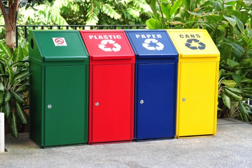 A row of four outdoor recycling bins positioned on a paved surface in front of lush green foliage, each designated for different waste types. The first bin on the left is green with a label indicating general waste, featuring a small icon and a no-smoking symbol. Next is a red bin marked for plastic recycling with a bold white recycling symbol and the word 'PLASTIC' in uppercase letters. Adjacent to it is a blue bin for paper recycling, also with a white recycling symbol and the word 'PAPER' prominently displayed. The last bin on the right is yellow, designated for cans, with a recycling symbol and the word 'CANS' in uppercase letters. Each bin has a round lid with a small keyhole lock, and they are evenly spaced in a straight line, situated against a backdrop of dense green leaves and plants, suggesting a location in a residential or commercial area in the UK, such as Staines. This setup exemplifies a typical waste separation station managed by companies like Commercial Waste Staines, providing environmentally responsible rubbish disposal solutions for local communities.
