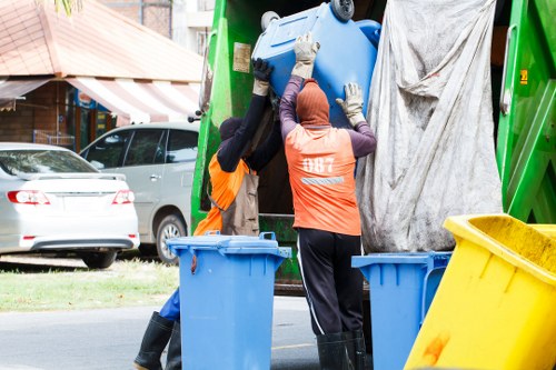 Two waste collection workers wearing high-visibility orange vests and gloves are lifting a large blue plastic recycling bin into the back of a green waste collection truck parked on a residential street in Staines. One worker is partially obscured, while the other faces away and uses both hands to steady the bin. The truck's rear door is open, revealing a protective grey cover partially draped over the opening. In the foreground, there are additional blue waste bins and a yellow waste container positioned on the pavement. Behind the scene, a row of parked cars, including a silver vehicle, can be seen alongside houses with brick walls, a striped awning, and a small front garden area, suggesting a typical suburban environment. The natural daylight and clear surroundings evoke a routine rubbish removal operation, typical for local commercial waste services like those offered by Commercial Waste Staines in the area near postcode TW18 and the town of Staines.