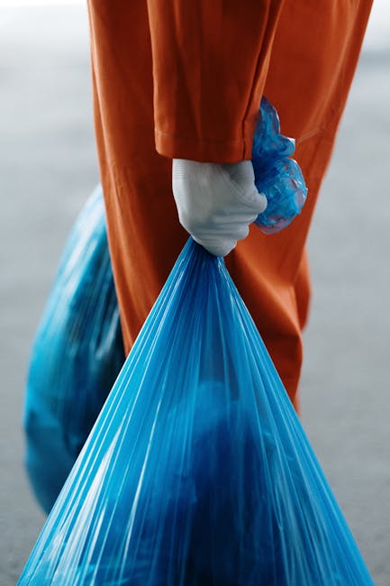 A large pile of mixed household waste is overflowing from multiple black, green, and red rubbish bins placed on a paved urban sidewalk, surrounded by scattered cardboard boxes, paper bags, plastic pac