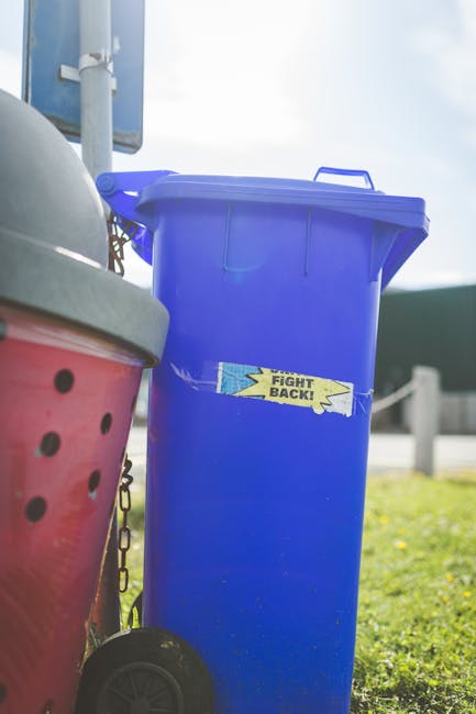 A group of five aerosol spray cans, each with different colours and labels, are positioned upright on a rough concrete surface in an urban outdoor setting. Behind the cans, there is a pile of mixed ru
