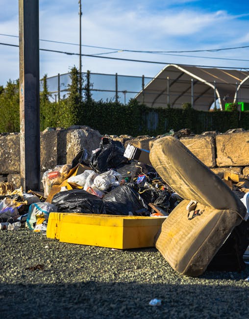 A pile of mixed domestic waste, including black and white plastic rubbish bags, a yellow plastic tray, and a dirty, worn car seat with visible fabric texture and stains, is situated on a gravel surfac