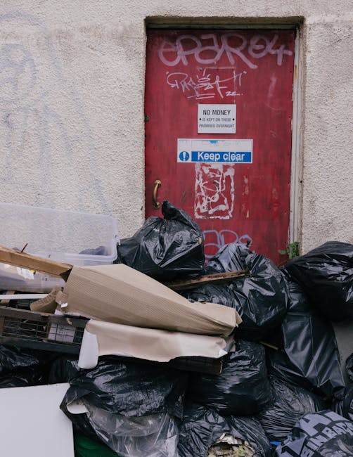 A pile of mixed domestic waste, including black and white plastic rubbish bags, a yellow plastic tray, and a dirty, worn car seat with visible fabric texture and stains, is situated on a gravel surfac